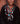 A close-up portrait of an elderly woman wearing multiple layered beaded necklaces and earrings. The jewelry features turquoise, coral-red, white, and black beads, with a prominent turquoise and black patterned pendant.