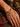 A close-up of an elderly person's weathered hand and wrist wearing a colorful braided prayer bracelet with a small circular pendant, set against a rustic background with brass prayer wheels and a patterned textile.