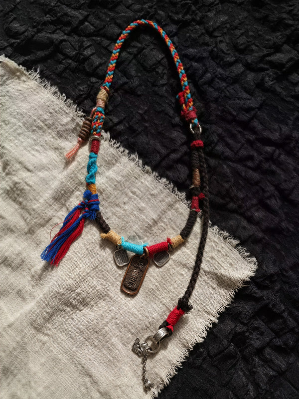 A handmade braided cord necklace with colorful tassels, wooden beads, metal charms, and a silver clasp, displayed on a split black and off-white textured fabric background.