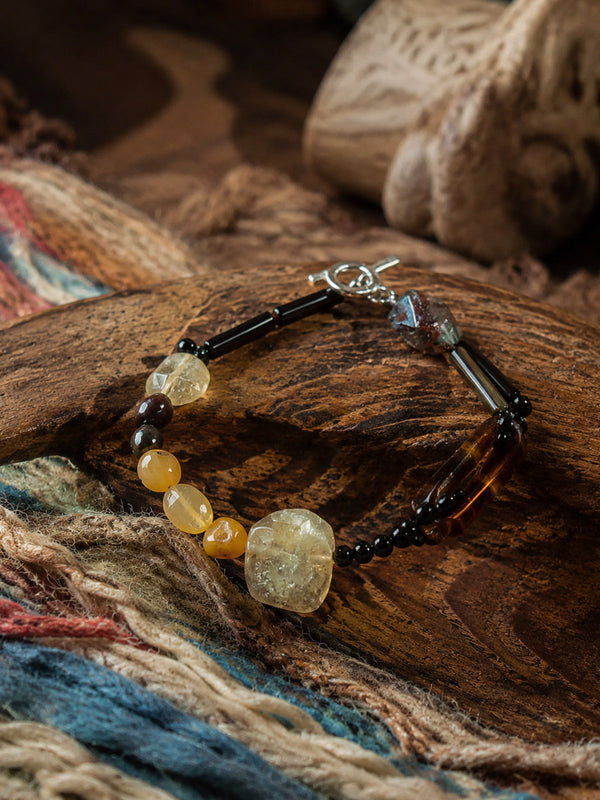 A beaded bracelet with black beads, amber-yellow beads, and a large faceted clear crystal pendant, displayed on a rustic wooden surface with a colorful woven textile underneath.