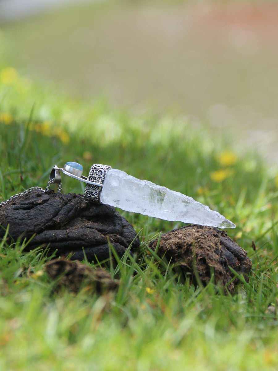 A clear crystal dagger pendant with a silver filigree hilt and a small light blue bead, resting on dark animal dung in a grassy outdoor setting.