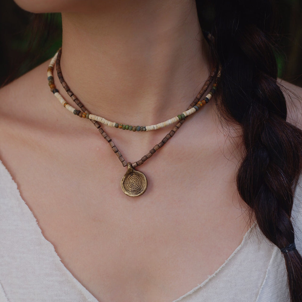 A close-up of a woman's neck and chest wearing a double-strand beaded necklace with a spiral pendant. The necklace features brown, cream, and green beads, with a bronze-toned spiral medallion pendant.