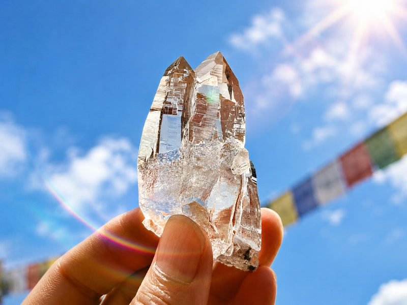 A hand holding a clear quartz crystal cluster against a bright blue sky with clouds and a blurred string of colorful prayer flags.