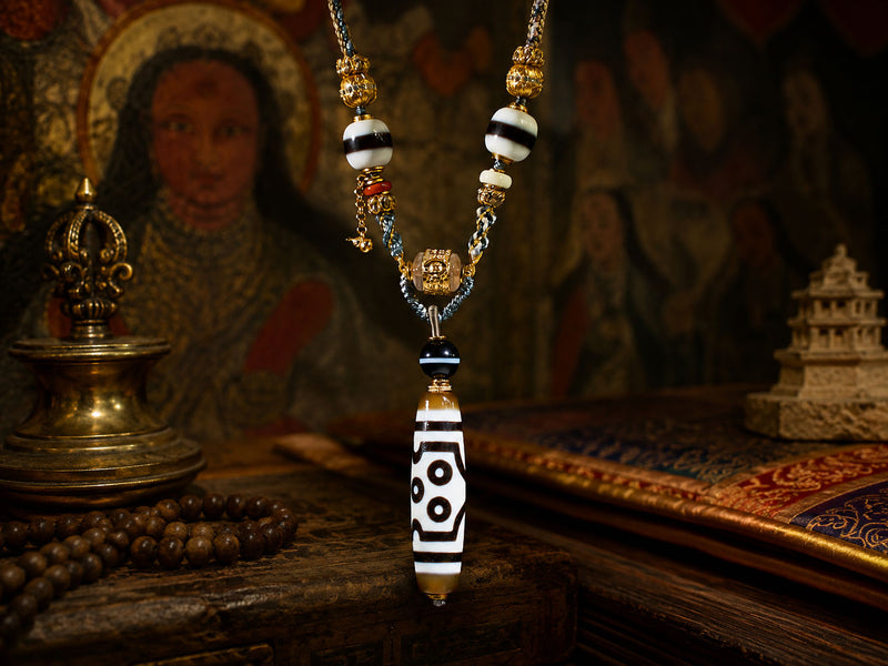 A beaded necklace with a long cylindrical pendant featuring black and white eye-like patterns, displayed on a wooden surface with a blurred religious icon and decorative objects in the background.