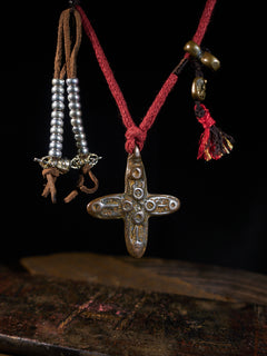 A red cord necklace featuring a central metallic cross pendant with circular details, accompanied by dangling beaded strands and tassels, displayed against a dark background.