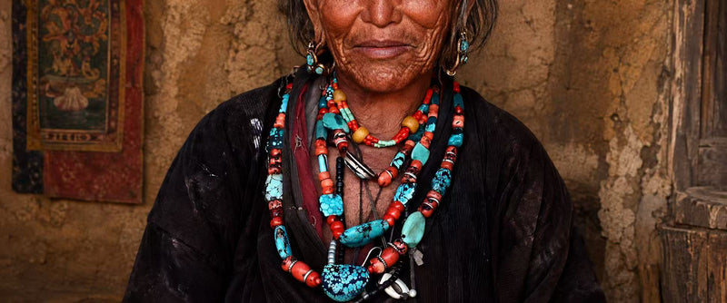 A close-up portrait of an elderly woman wearing multiple layered beaded necklaces and earrings in turquoise, coral, red, and white, set against a rustic textured wall.