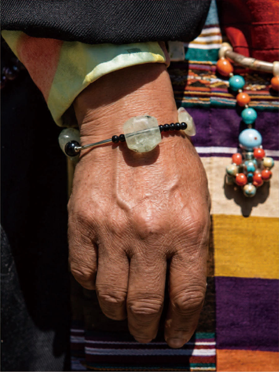 A close-up of a person's hand wearing a beaded bracelet with a large light green stone and black beads, set against a colorful striped textile background.
