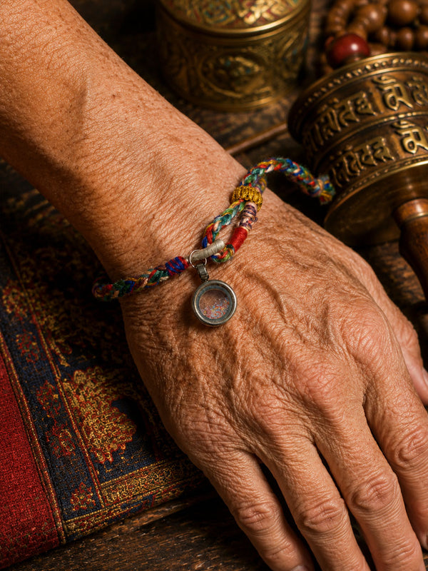 A close-up of an elderly person's weathered hand and wrist wearing a colorful braided prayer bracelet with a small circular pendant, set against a rustic background with brass prayer wheels and a patterned textile.