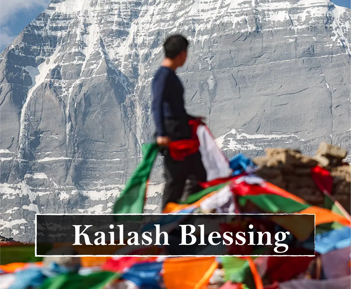 A scenic photograph of a snow-covered mountain with a blurred person standing in front of it, and colorful prayer flags in the foreground. A text banner across the lower portion reads 'Kailash Blessing'.