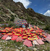 A large pile of red prayer flags and colorful painted fabric banners spread across the foreground, set against a rocky mountain slope under a blue sky with white clouds.