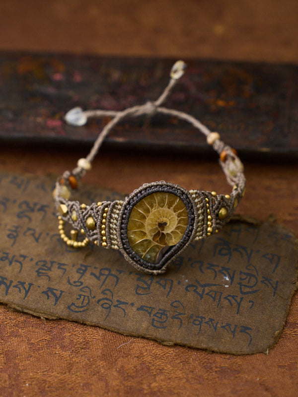 A handmade macrame bracelet featuring a central fossilized ammonite shell specimen, displayed on a textured brown surface with Tibetan script.