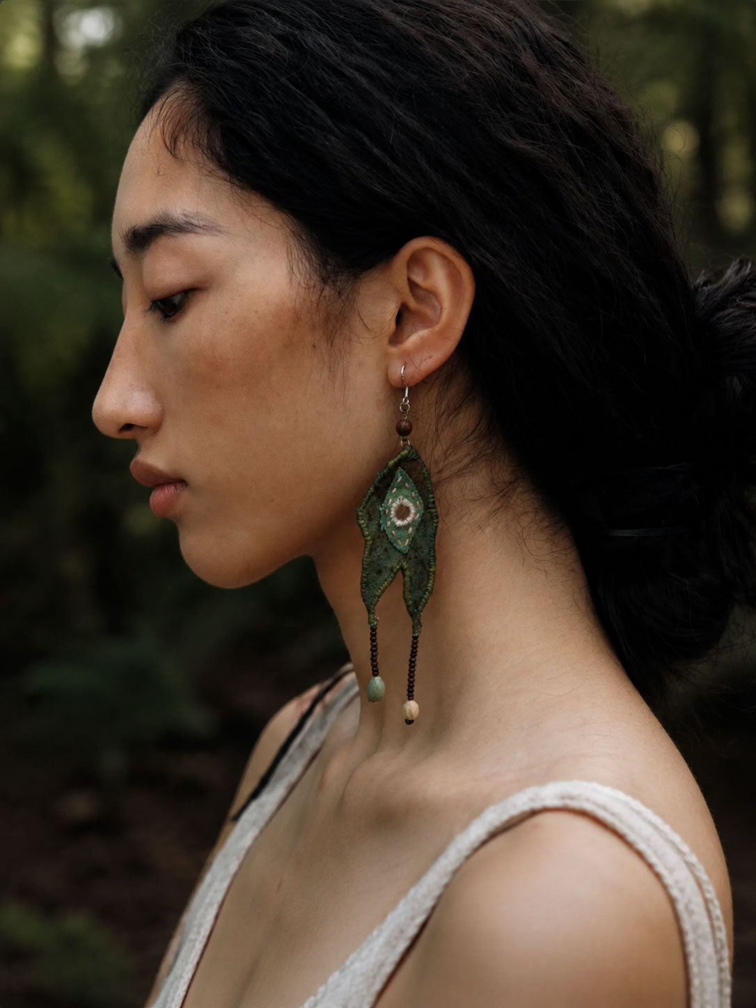 A woman in profile wearing a long, ornate green dangling earring with bead details, photographed outdoors against a blurred forest background.