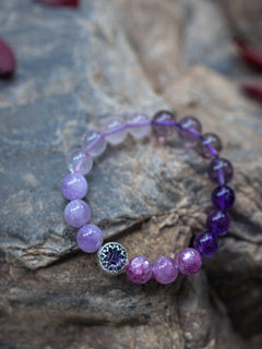 A beaded stretch bracelet featuring purple and lavender beads, a silver-toned bezel-set blue gemstone accent, and a dark purple amethyst end, displayed on a textured gray stone surface.