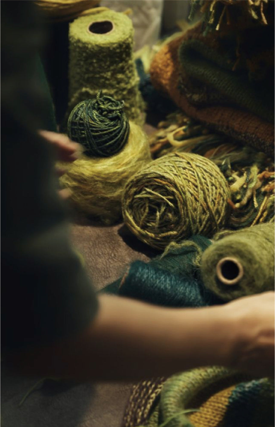 A close-up scene of multiple skeins and cones of yarn in olive green, dark green, and yellow-green tones, with a person's hands and arm visible handling the fibers in a dimly lit setting.
