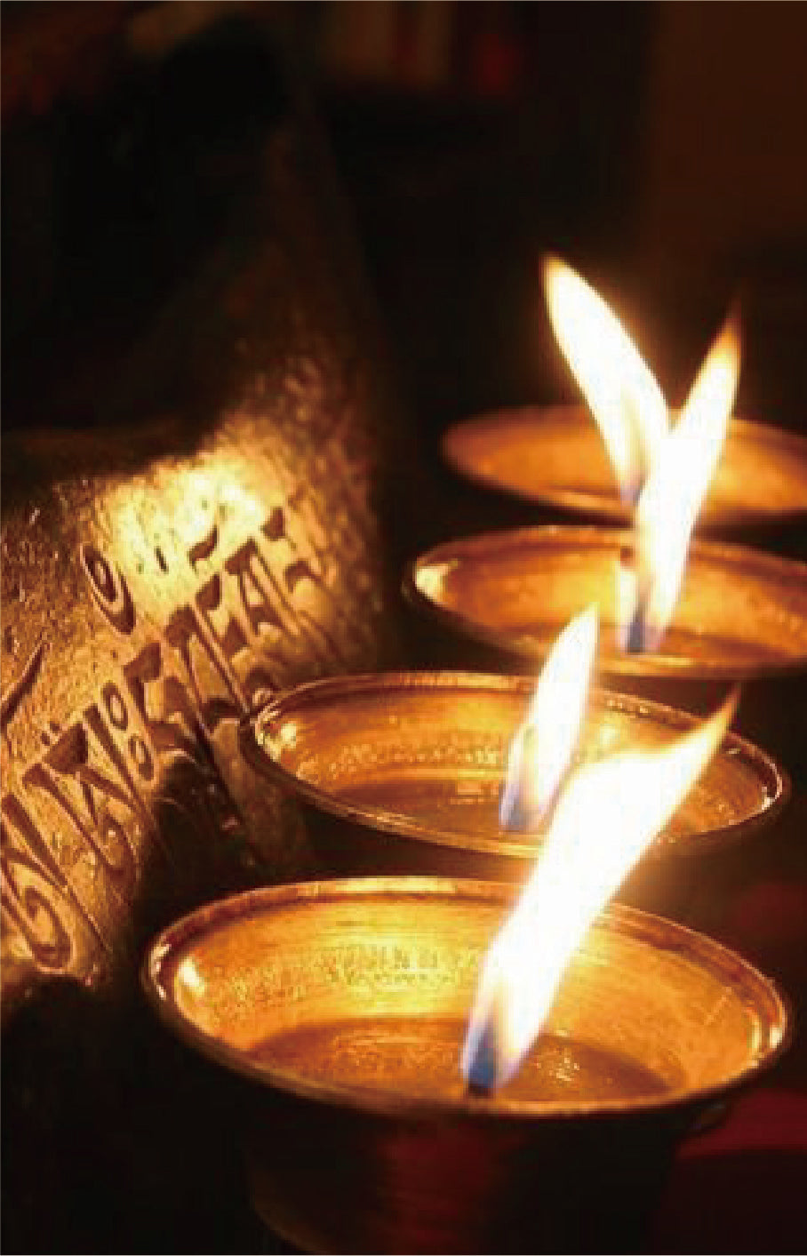 A close-up shot of several small brass oil lamps (diyas) with bright yellow and blue flames burning, set against a dark background with a metallic engraved object on the left.