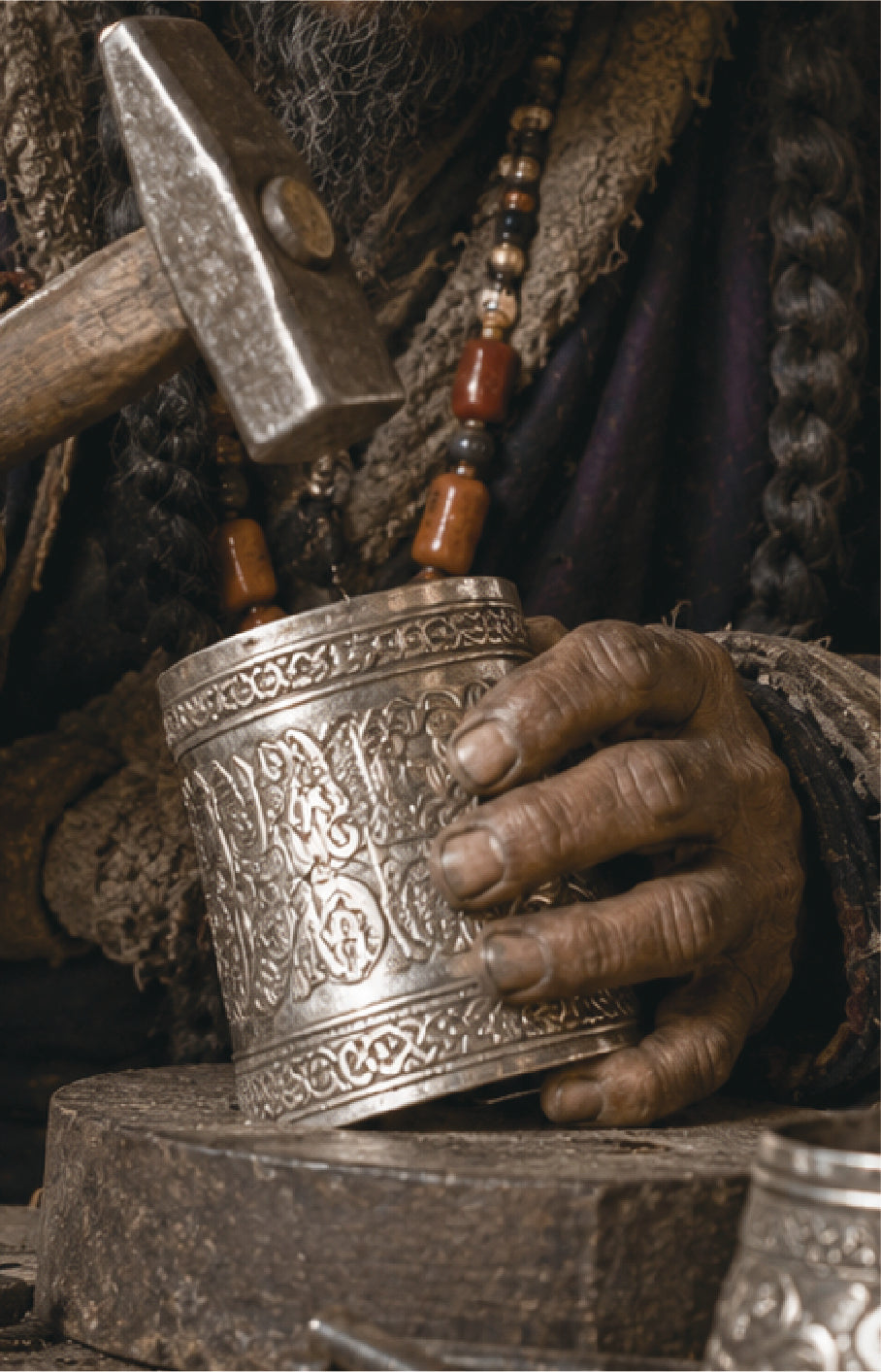 A close-up of a hand holding an ornate engraved silver metal cup or tumbler, with a hammer resting nearby and a beaded necklace visible against dark clothing in a rustic, handcrafted setting.