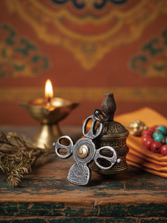 A silver-toned Tibetan wind spinner with a central windmill design and a small wind chime, displayed on a rustic wooden surface next to a burning brass oil lamp and other decorative elements.