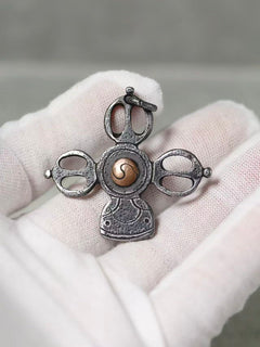 A small ornate pendant or charm with a silver-toned metal design and a central gold-toned spiral detail, displayed on a white gloved hand against a soft gray background.