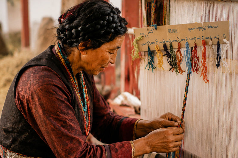 A woman in traditional clothing works on a handloom, weaving a long, narrow textile with colorful thread tassels hanging on a wooden board behind her.