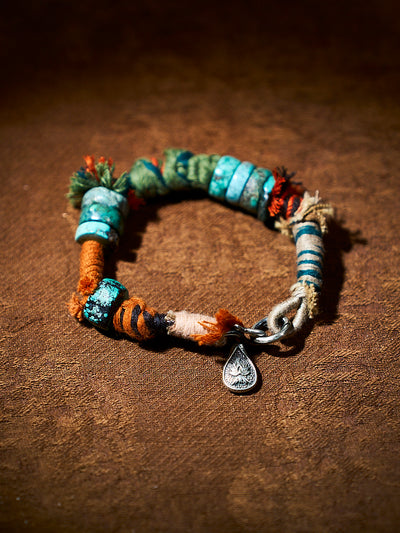 A handmade beaded bracelet with turquoise, green, orange, and brown beads, a silver chain, and a teardrop-shaped silver charm, displayed on a textured brown leather background.