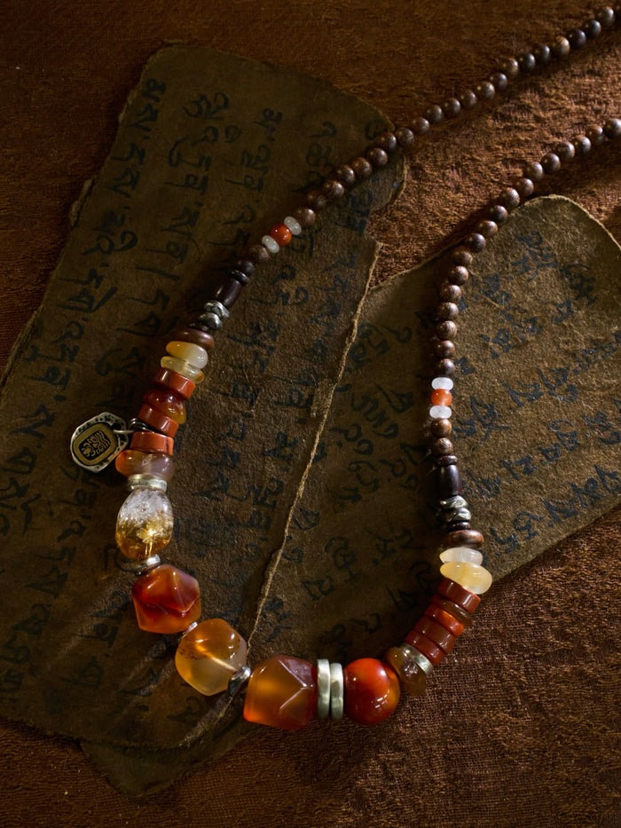 A beaded necklace with a mix of dark brown wooden beads, translucent orange and red agate stones, clear crystal beads, and small silver spacer rings, displayed on a textured brown leather background with an antique book.